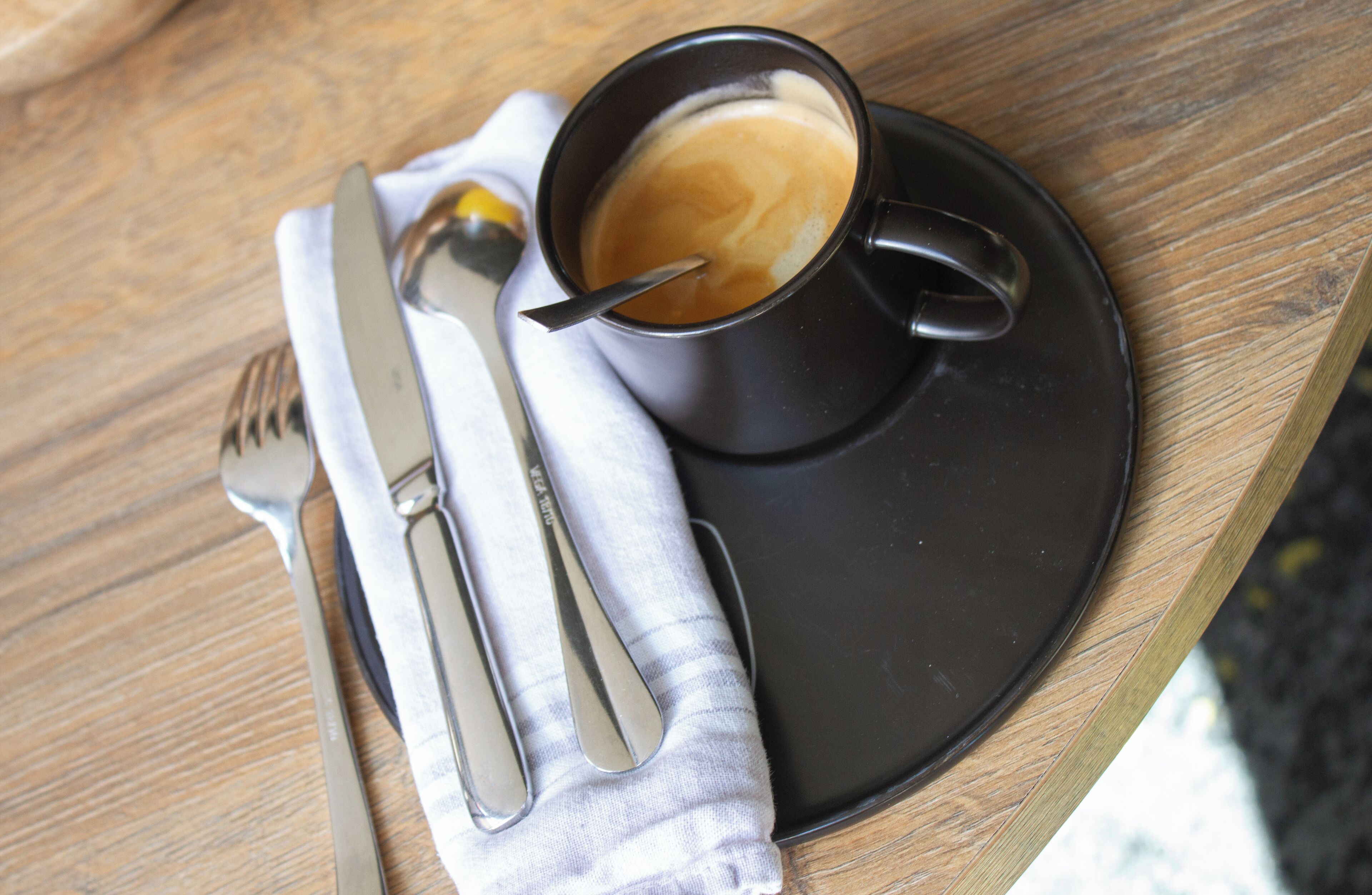 Una taza de café sobre un platito negro con una cuchara y un tenedor sobre una servilleta blanca, colocada sobre una mesa de madera.