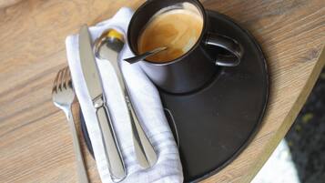 Una taza de café sobre un platito negro con una cuchara y un tenedor sobre una servilleta blanca, colocada sobre una mesa de madera.