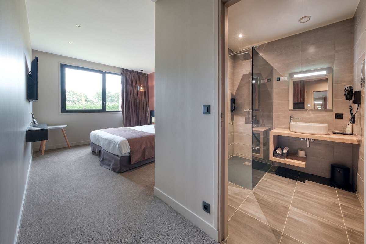 A modern hotel room with a large bed, a desk, a TV, a bathroom with a glass shower, and a large window.