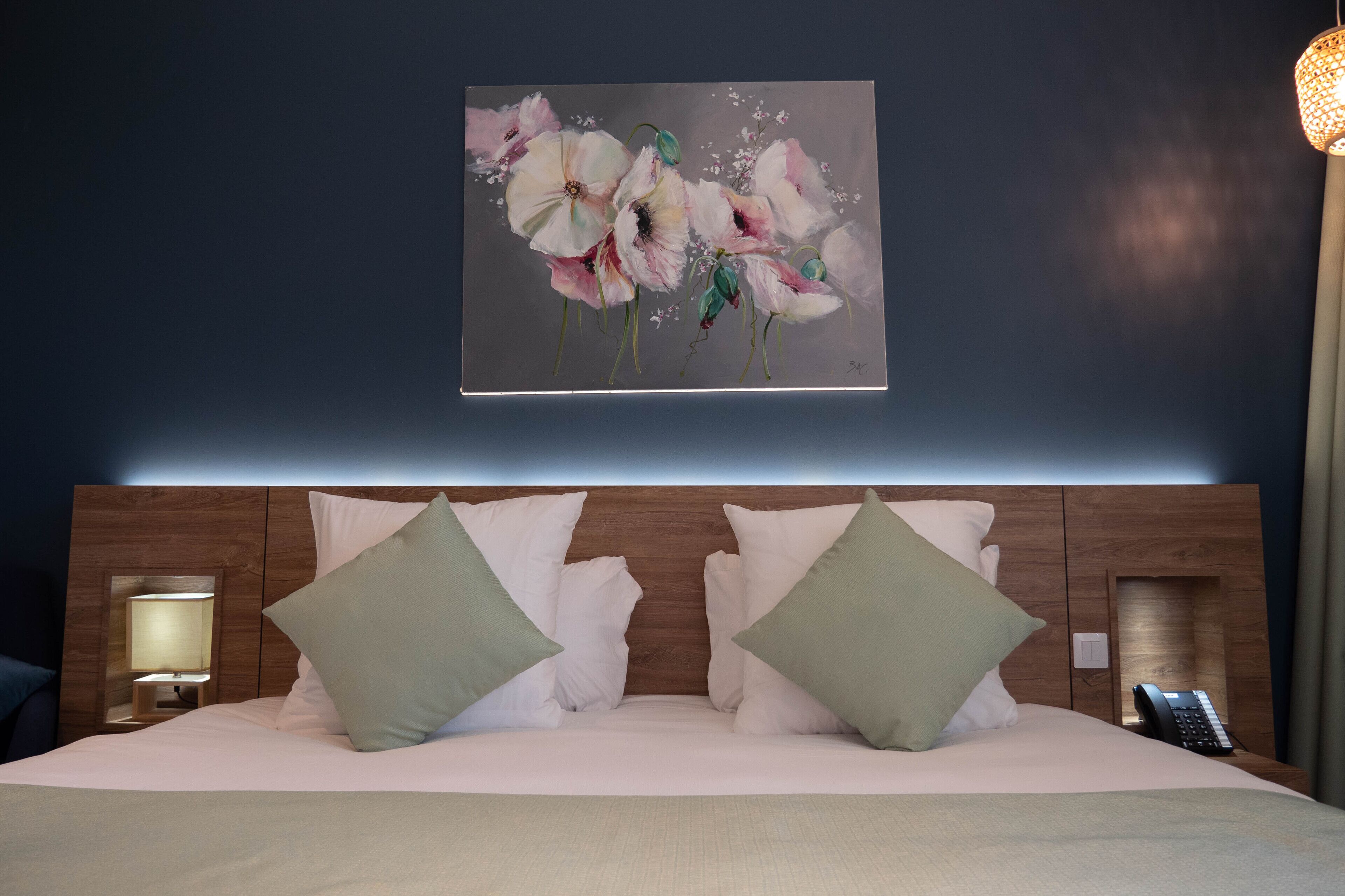 A neatly made bed with white and green pillows, a wooden headboard, and a painting of flowers on the wall.