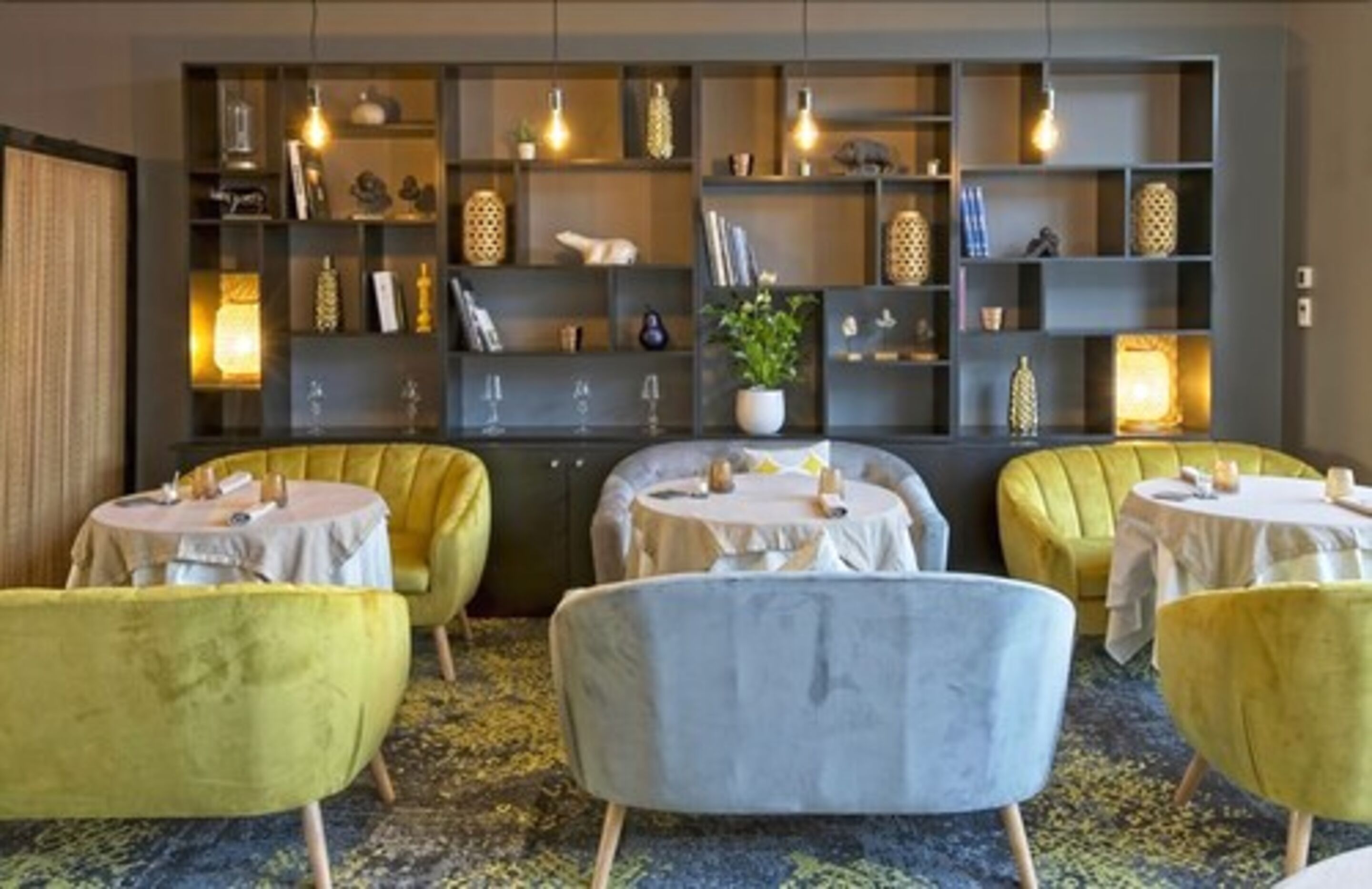 A hotel room with a seating area featuring round tables and yellow chairs, a bookshelf with decorative items, and a potted plant.