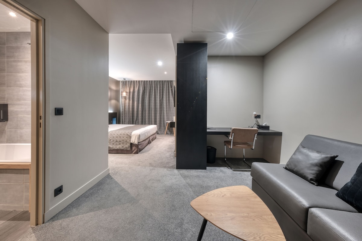 A modern hotel room with a grey sofa, a wooden coffee table, a desk, and a bed with a grey headboard.