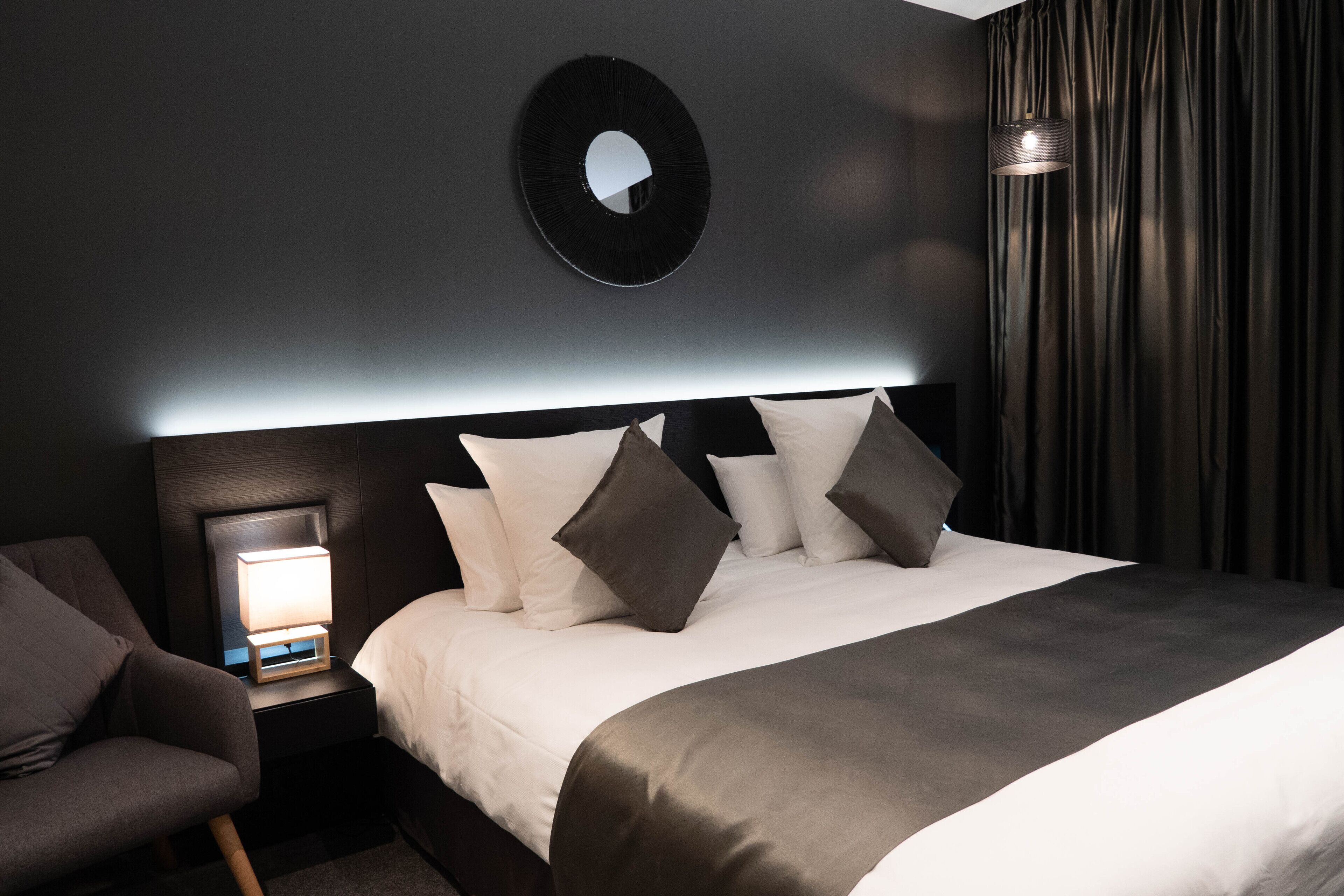 Ein modernes Hotelzimmer mit einem ordentlich bezogenen Bett, einer Nachttischlampe, einem runden Spiegel und einem dunklen Vorhang.