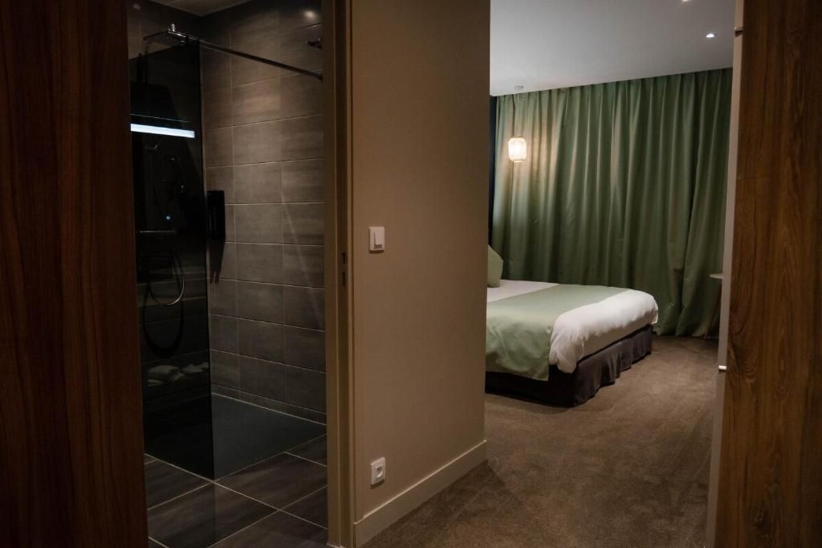 Ein Hotelzimmer mit einer gläsernen Duschkabine, einem Bett mit grünem Vorhang und Teppichboden.