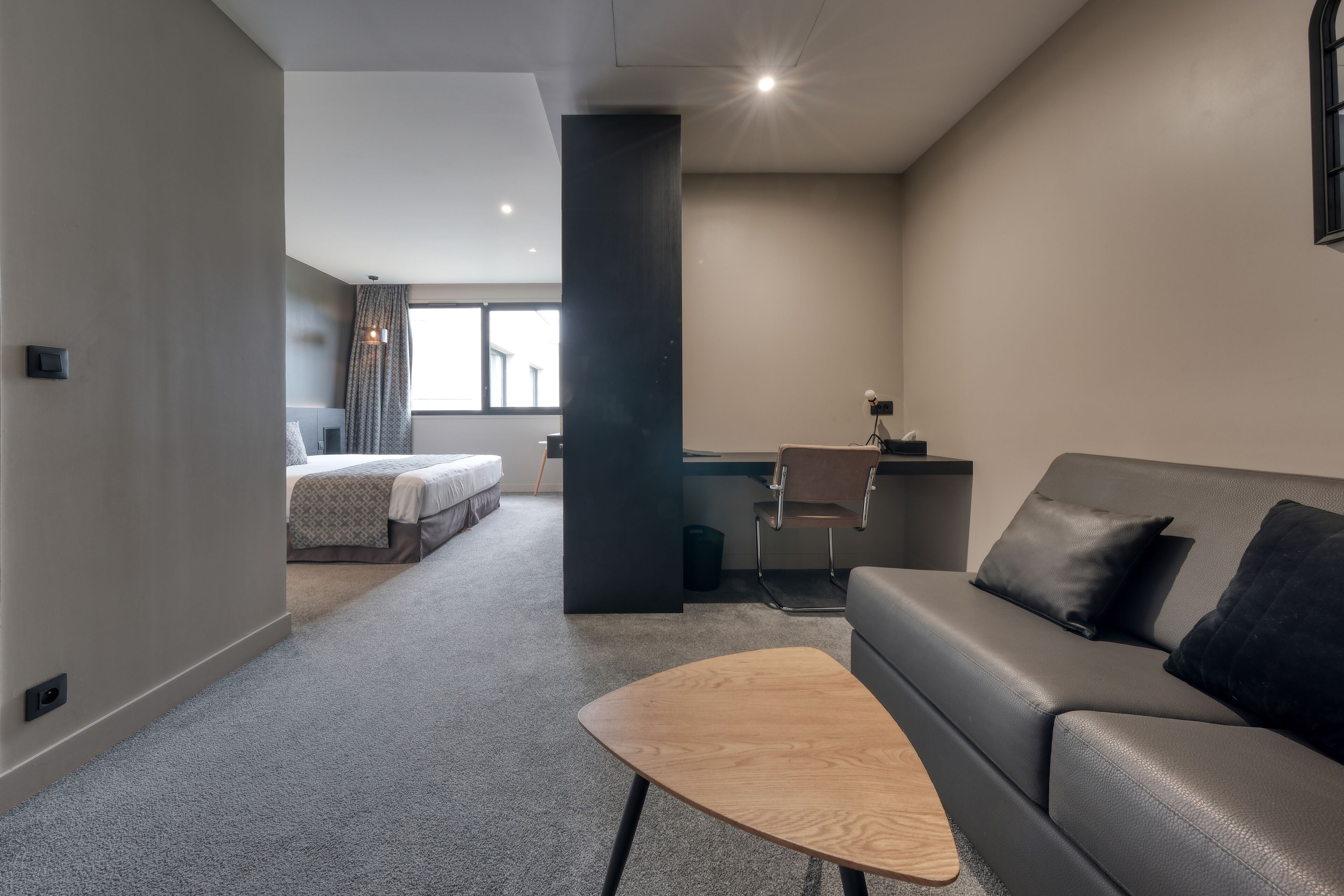 Een moderne hotelkamer met een grijze bank, een houten salontafel, een bureau met stoel, en een bed met een beddengoed met een patroon.