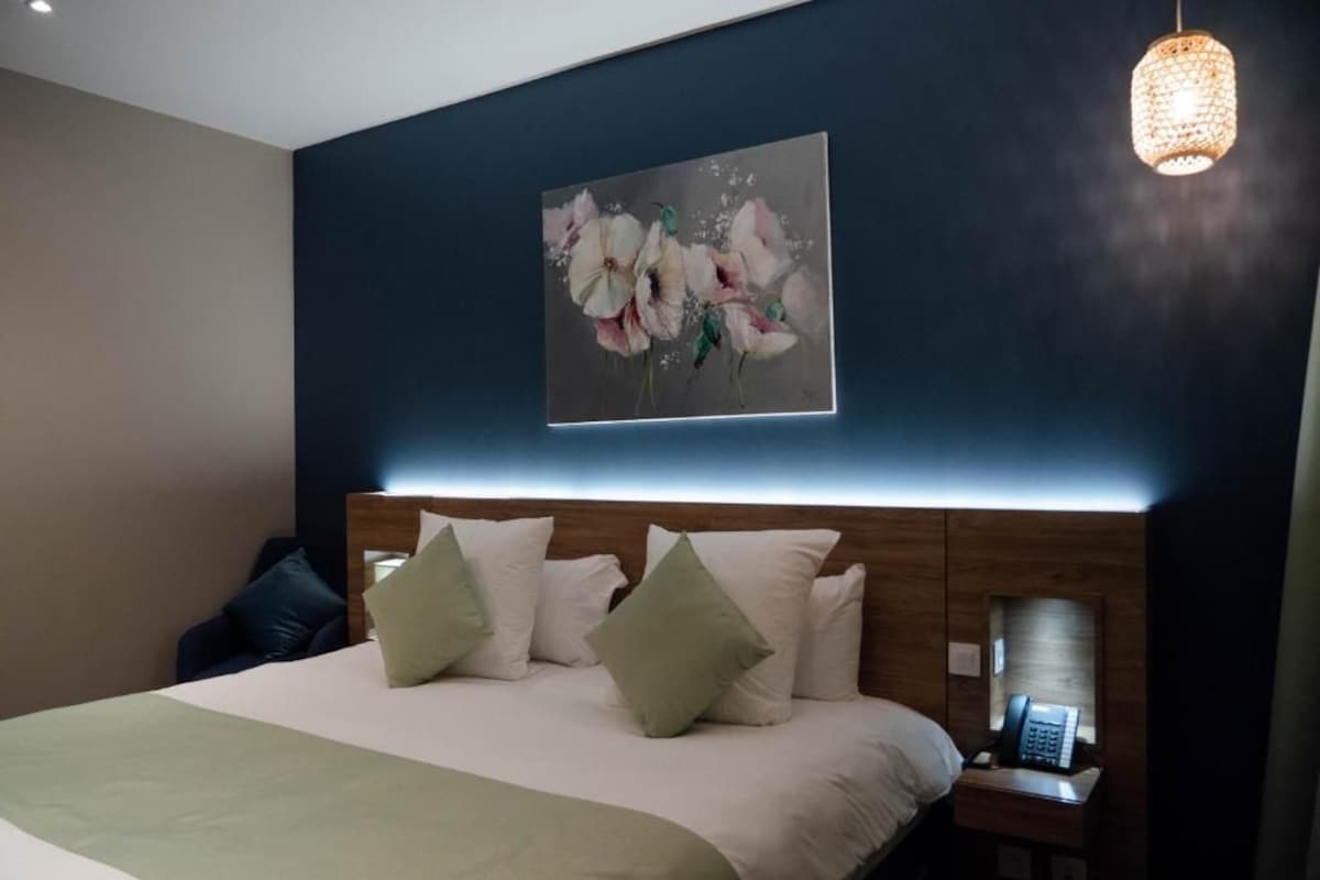 A modern hotel room with a large bed, a wooden headboard, a bedside table with a phone, and a wall-mounted painting of flowers.