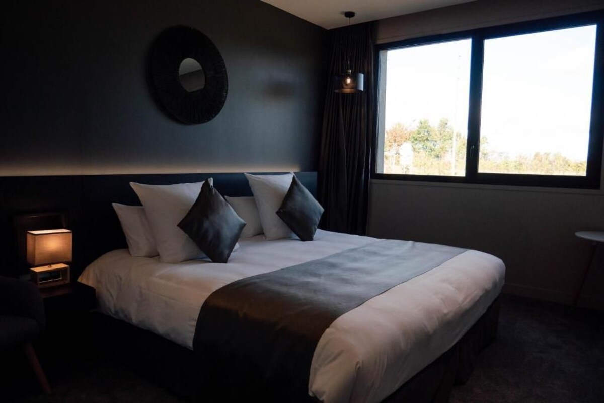 A hotel room with a large bed, a bedside lamp, a round mirror on the wall, and a window with curtains.