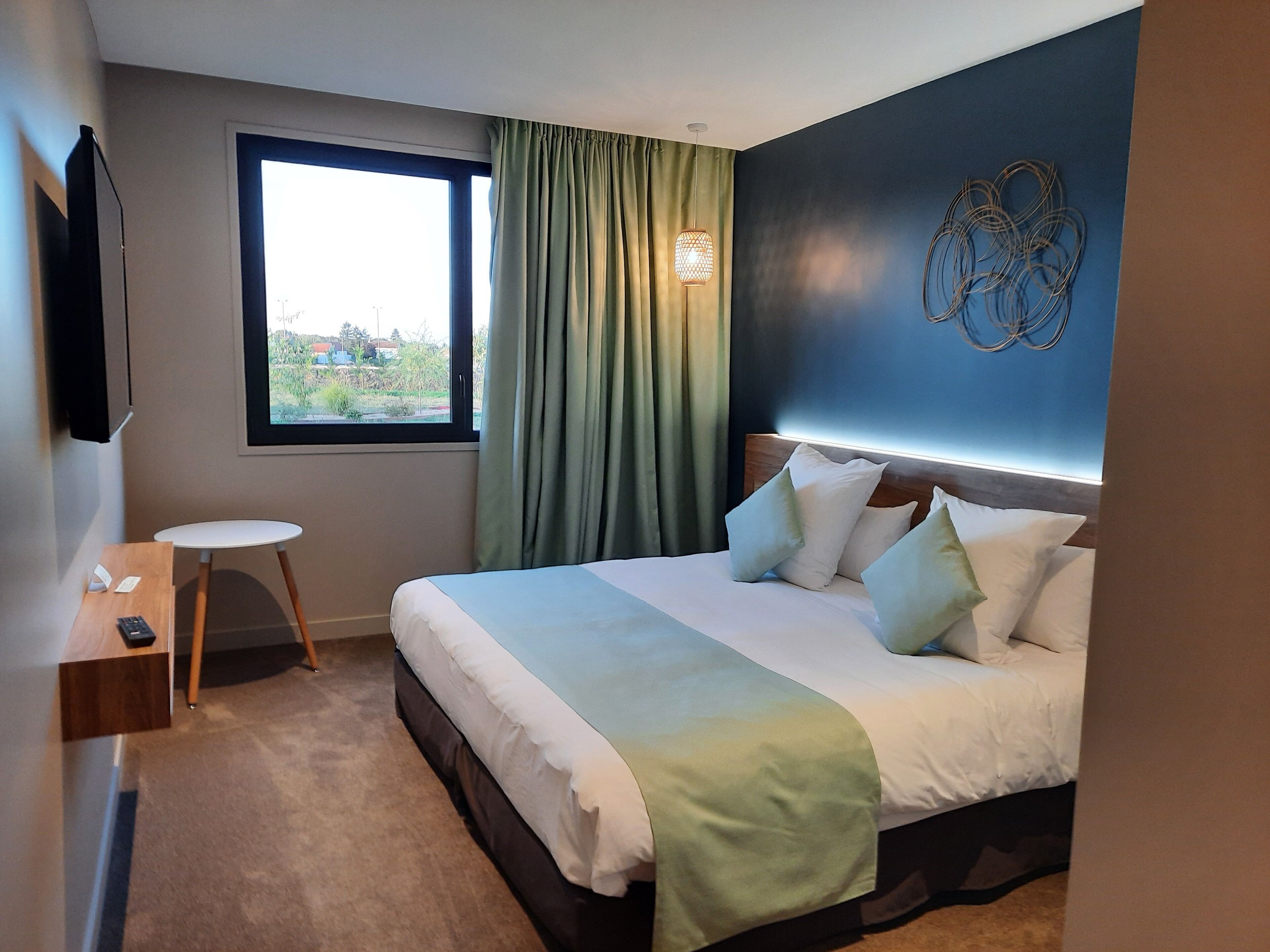 Ein Hotelzimmer mit einem Bett, einem kleinen Tisch, einem Stuhl, einem Fenster mit Vorhängen und einer Wandleuchte.