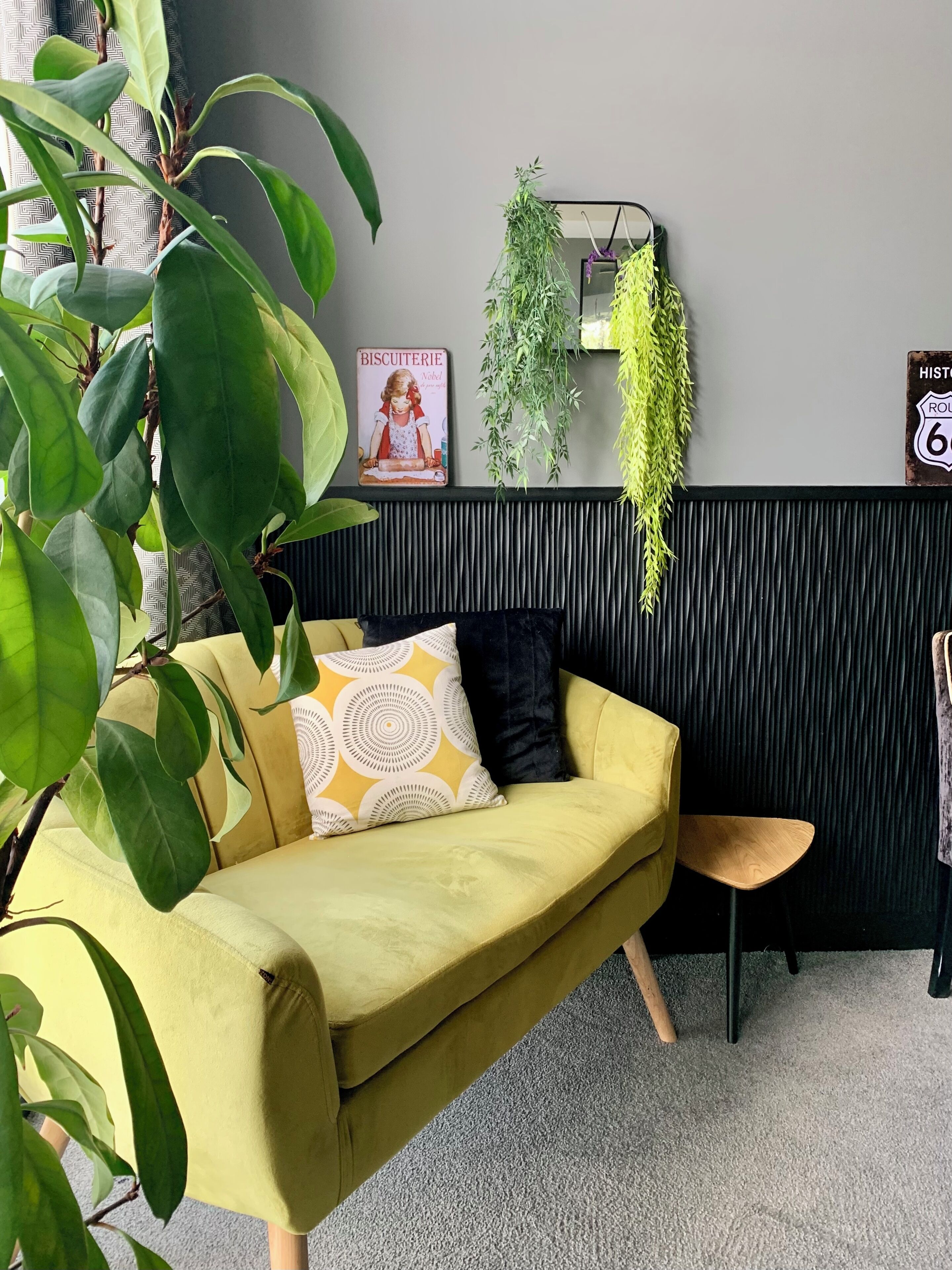 A yellow sofa with patterned pillows, a small wooden stool, and a large green plant in the foreground.