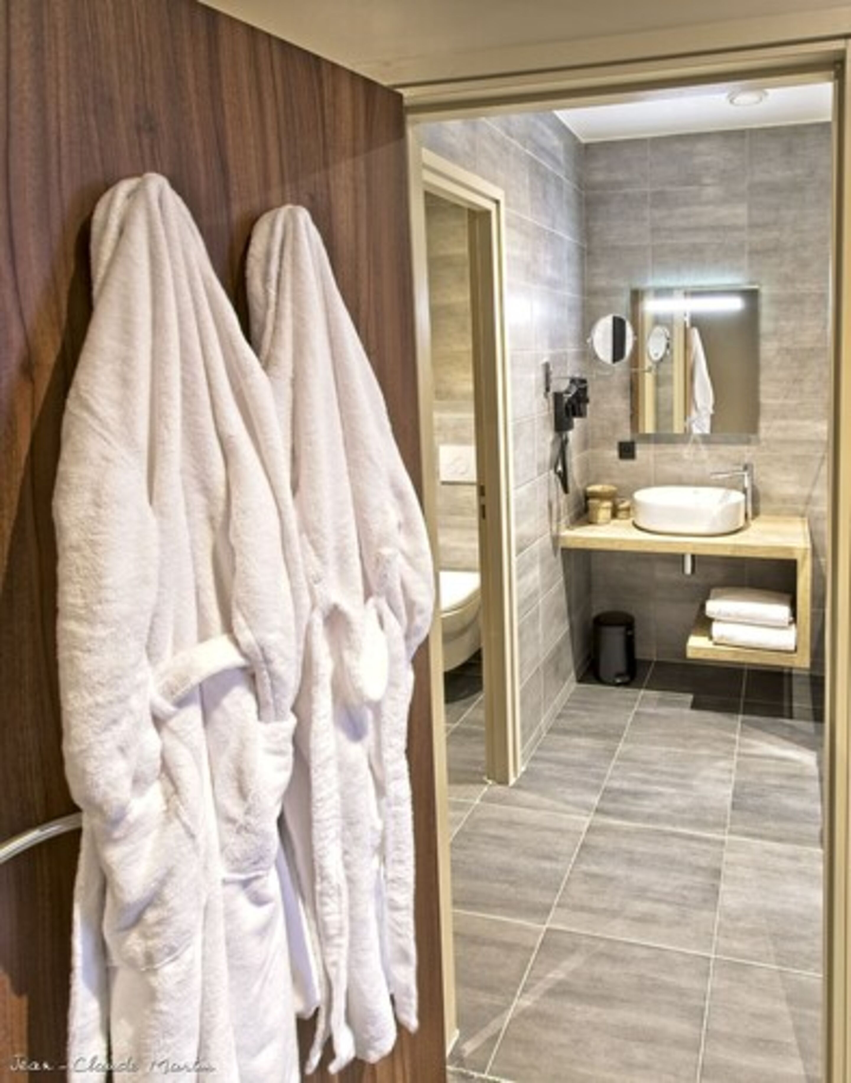 A modern bathroom with a wooden vanity, a round mirror, and a towel rack with two white bathrobes.