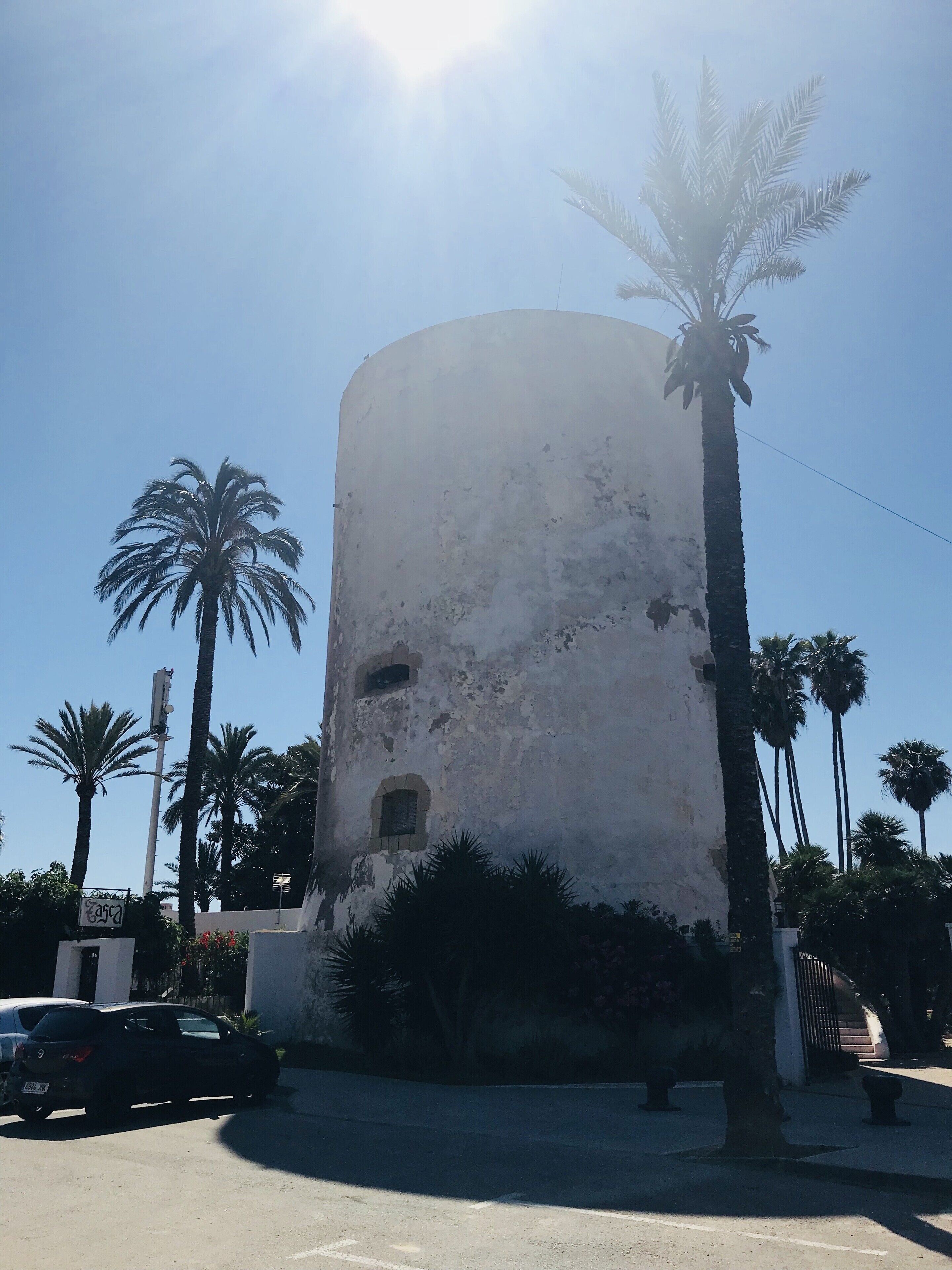 A cylindrical building with a weathered facade, surrounded by palm trees and a parked car.