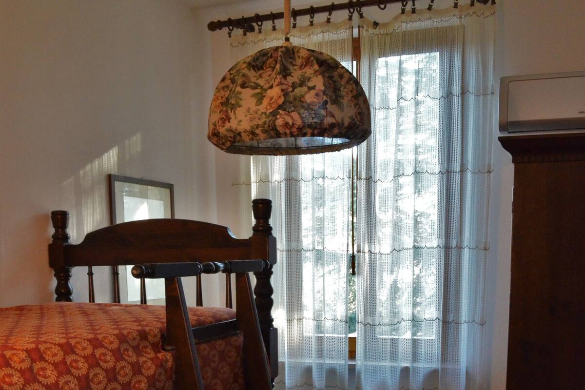 Een kamer met een stapelbed, een houten dressoir, een raam met dunne gordijnen en een hanglamp.