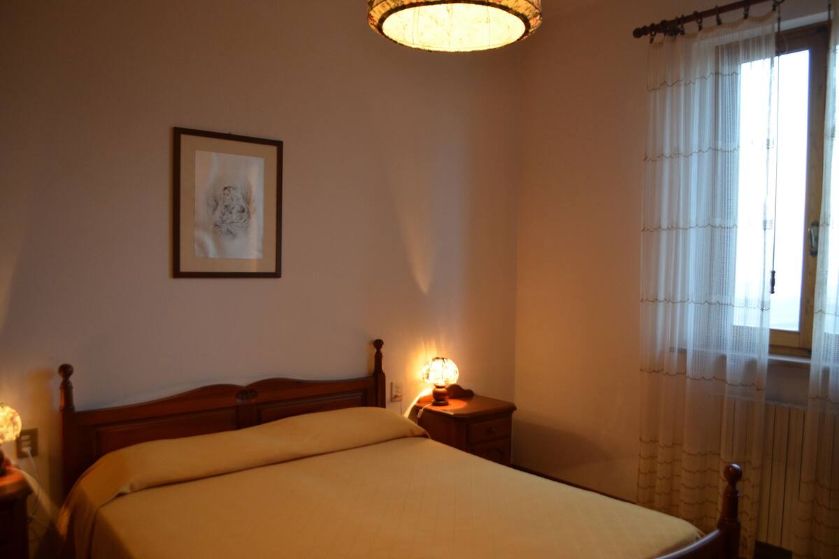 Een slaapkamer met een houten bed, een bedlampje, een ingelijste foto aan de muur en een raam met dunne gordijnen.