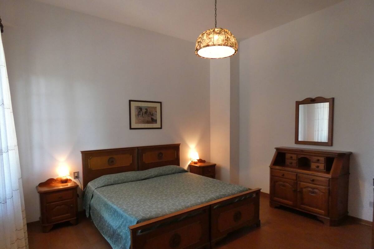 Een slaapkamer met een houten bed, nachtkastjes met lampen, een kaptafel, een hanglamp, een ingelijste foto aan de muur en een spiegel.