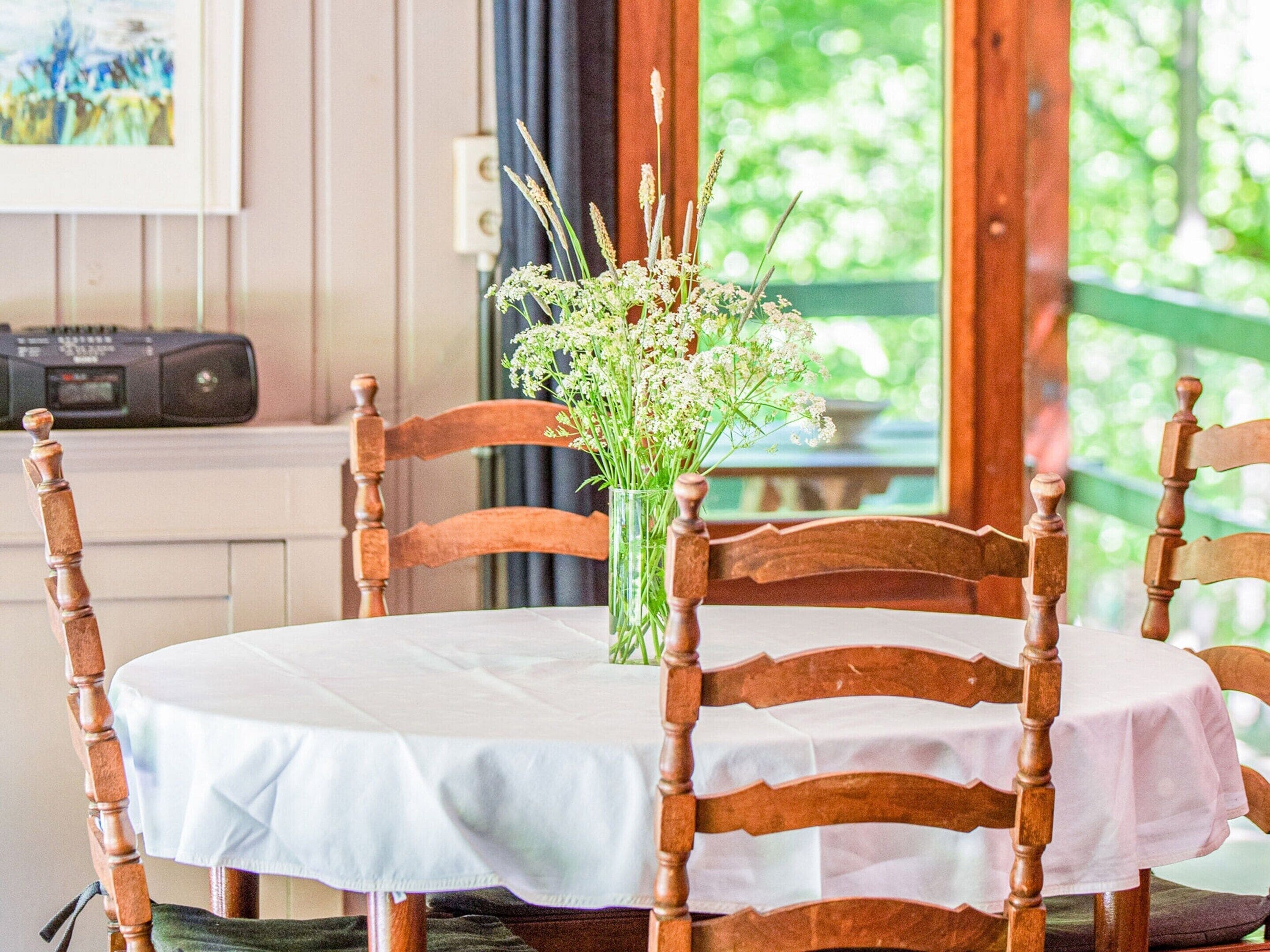 Une table ronde en bois, recouverte d’une nappe blanche, est entourée de chaises en bois dont les pieds sont ornés de motifs tournés. Un vase rempli de fleurs blanches est posé sur la table.