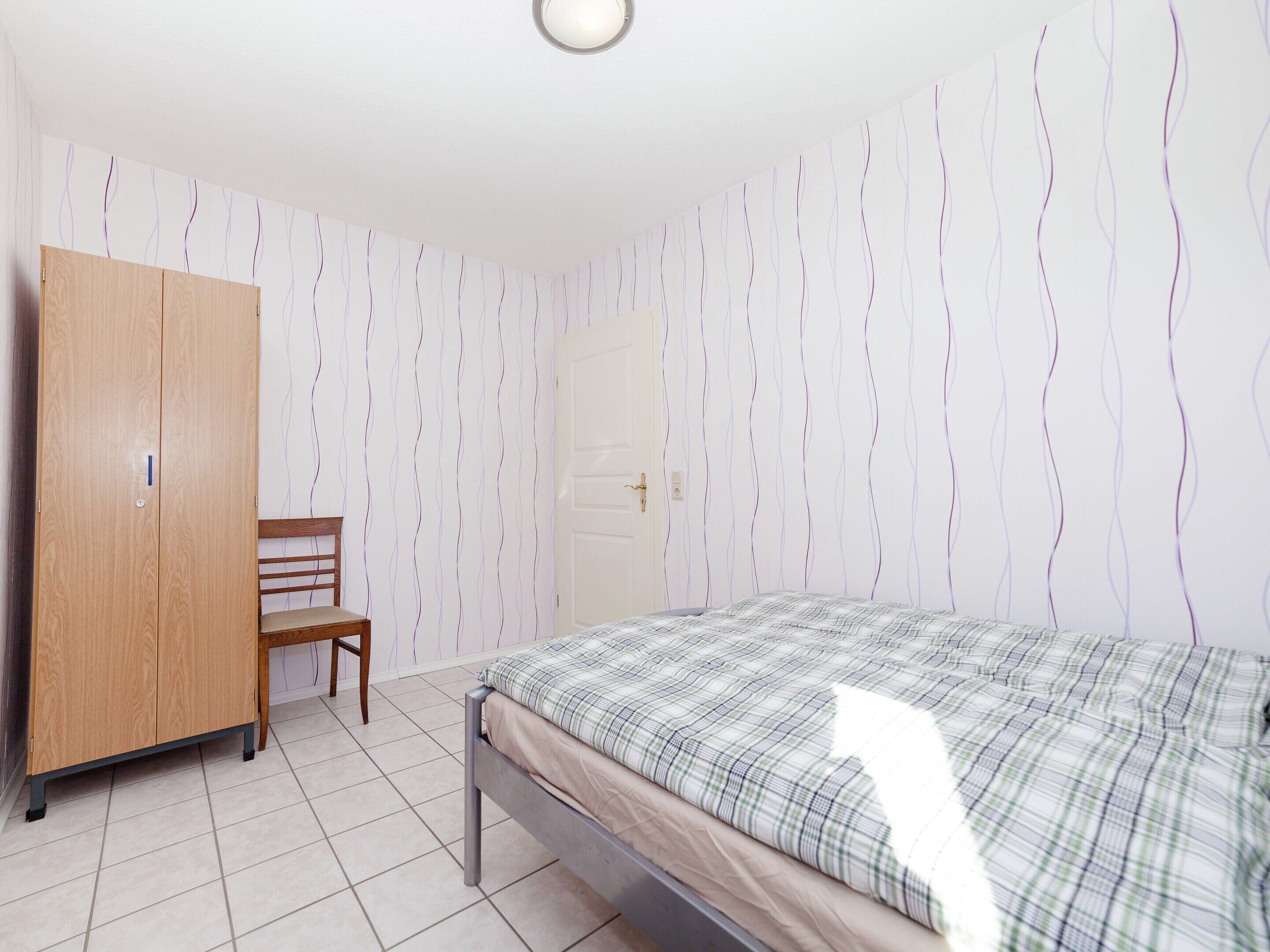 A bedroom with a bed, a wooden wardrobe, a chair, and a door.