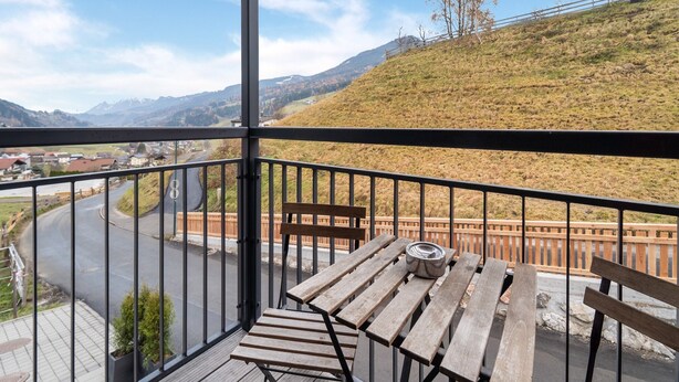 A balcony with a wooden table and chairs, overlooking a winding road and a mountainous landscape. - Apartment in St. Georgen (Bruck an der Grossglocknerstrasse)