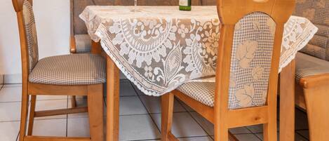 Une table à manger recouverte d’une nappe en dentelle, entourée de chaises en bois avec des coussins à carreaux. Une bouteille de vin est posée sur la table.