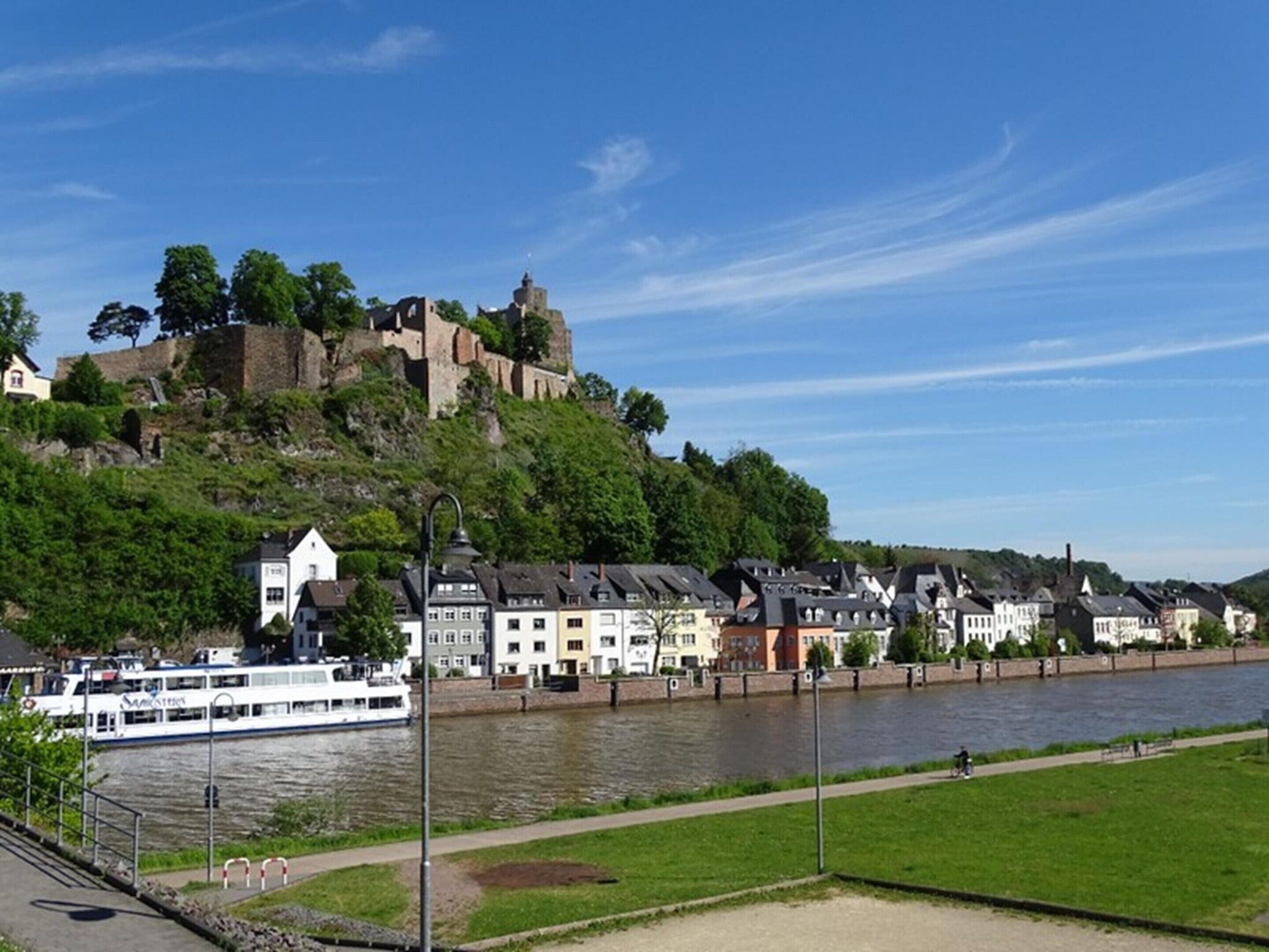 A riverside town with historic buildings, a castle on a hill, and a boat docked along the river.