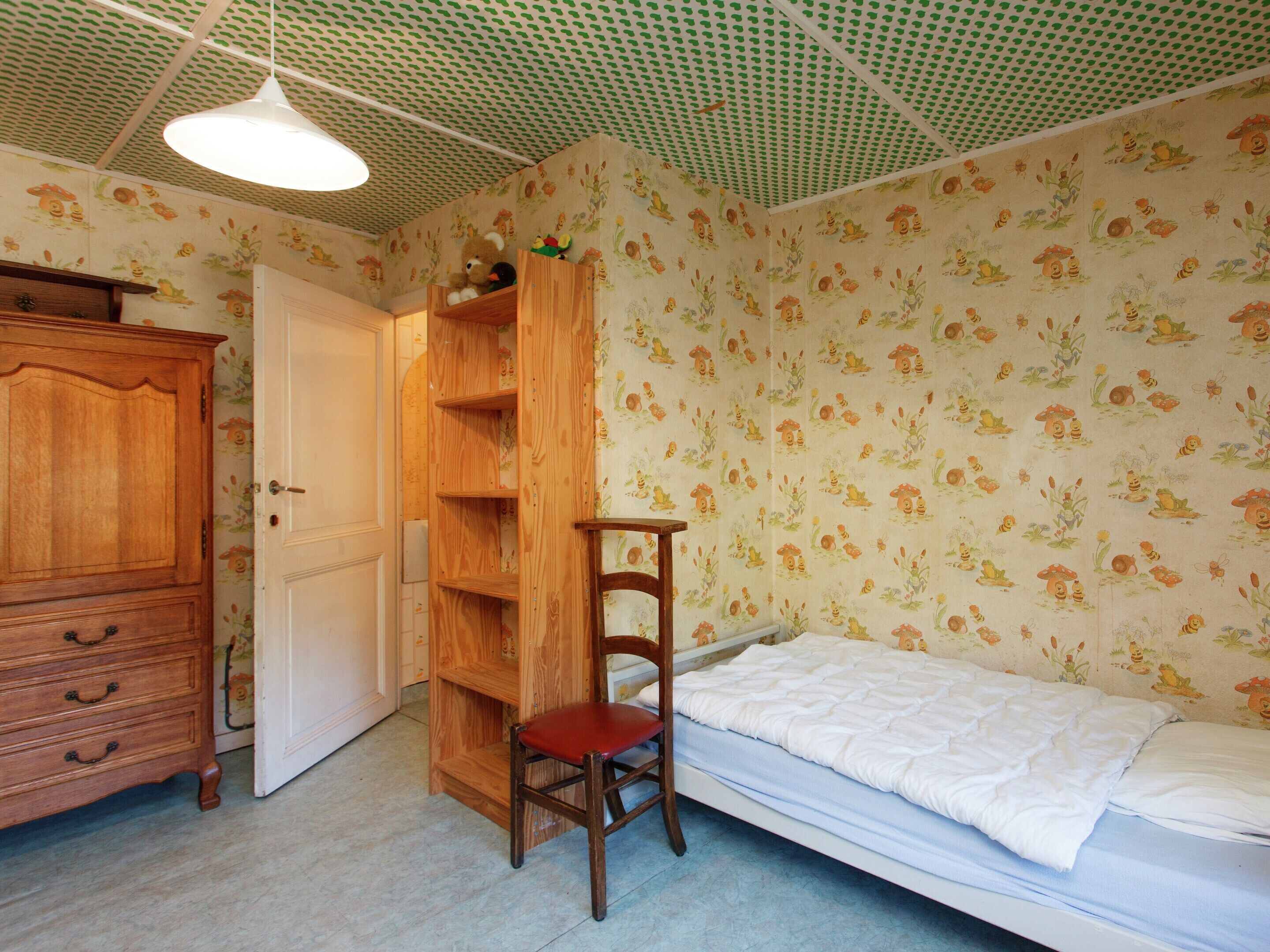 A bedroom with a wooden wardrobe, a built-in bookshelf, a single bed, and a chair.
