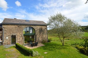 A stone building with a large arched window, a patio with outdoor seating, and a tree in the foreground. - Gite With Lake View (Bastogne)