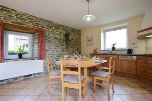 A kitchen with a stone wall, wooden dining table, chairs, and a window with curtains. - Gite With Lake View (Bastogne)