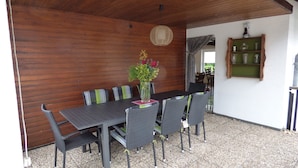An outdoor dining area with a wooden ceiling, a table with chairs, a vase with flowers, and a shelf with glassware. - Geraumige Ferienwohnung im Sauerland (Olsberg)