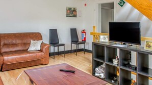 Een woonkamer met een bruine leren bank, twee zwarte stoelen, een houten salontafel, een televisie op een meubel en een brandblusser aan de muur.