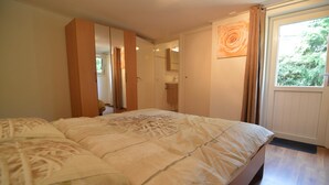 A bedroom with a large bed, a wardrobe, a bathroom with a mirror, and a view of greenery through a window.