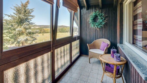 A balcony with a wicker chair, a small round table with purple containers, and a view of trees and hills. - Trierscheid Countryside Escape (Trierscheid)