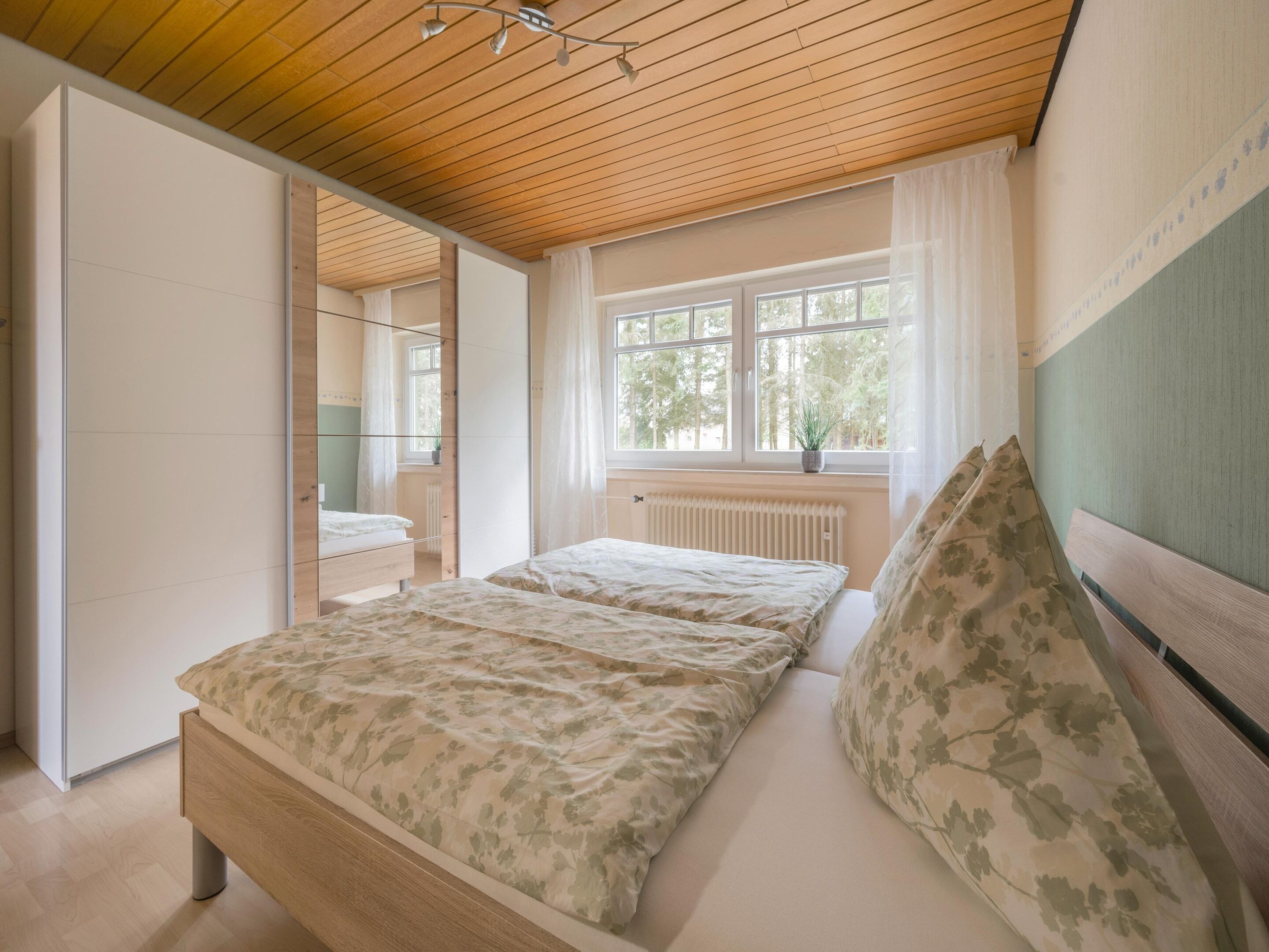 Un dormitorio con techo de madera, cama con ropa de cama floral, ventana con vistas a árboles y un radiador.