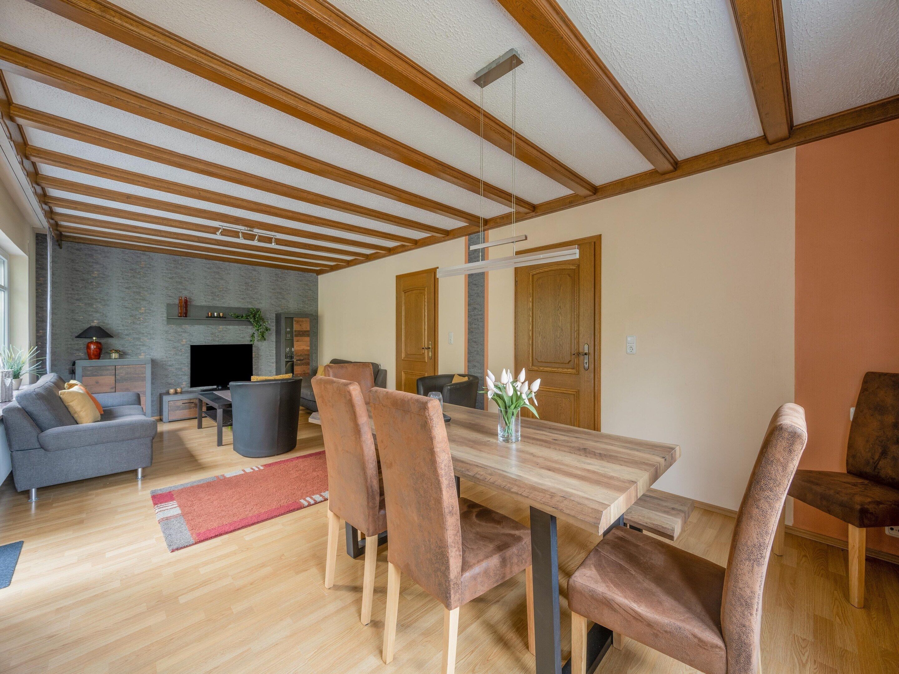 A spacious living area with a wooden ceiling, a dining table with chairs, a sofa, and a TV.