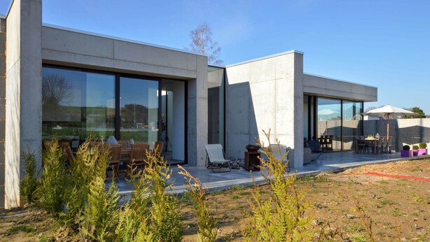 Une maison moderne dotée de grandes baies vitrées, d’un patio avec mobilier d’extérieur et offrant une vue sur le paysage environnant.