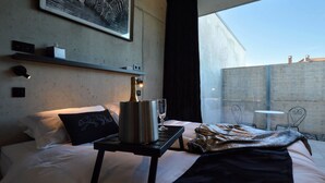 Une chambre d’hôtel moderne avec un lit, une table de chevet sur laquelle se trouve une bouteille de champagne, un verre et une couverture. Une grande fenêtre offre une vue sur un immeuble et un mur en béton.
