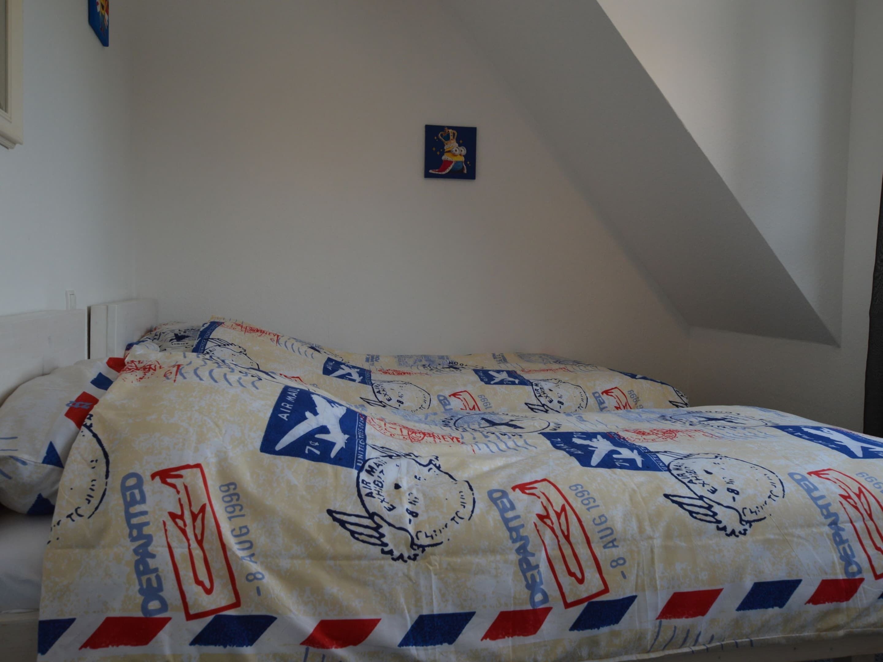 Un letto con una coperta decorata con motivi postali, una piccola cornice con un'immagine appesa al muro e un soffitto spiovente.