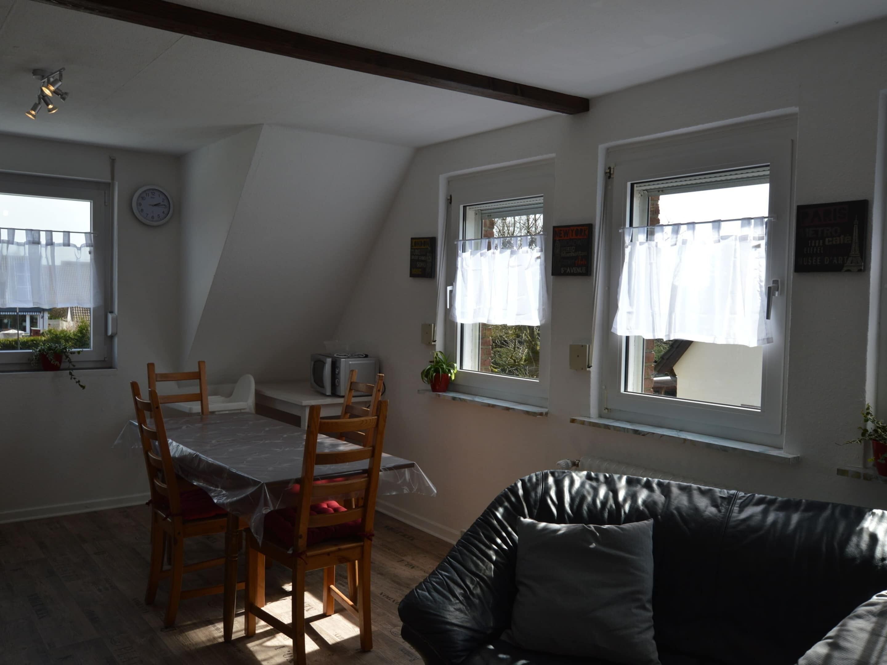 A dining area with a table and chairs, a black leather couch, and two windows with curtains.