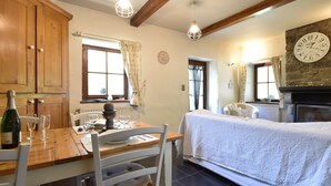 Une pièce chaleureuse avec une table à manger en bois, des chaises, un lit recouvert d’une courtepointe blanche, une cheminée en pierre et une grande horloge murale.