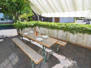 An outdoor wooden table with benches, set up under a white canopy, surrounded by greenery and a white wall.