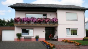 A two-story house with a balcony adorned with pink flowers, a front door, and a small garden area with colorful flowers. - Cozy Apartment in Strotzbüsch near Eifel Center (Strotzbusch)