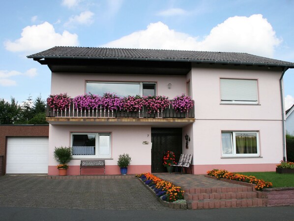 A two-story house with a balcony adorned with pink flowers, a front door, and a small garden area with colorful flowers.