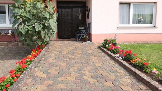 A paved pathway leading to a building with a dark entrance door, a small outdoor seating area, and flower beds on either side. - Cozy Apartment in Strotzbüsch near Eifel Center (Strotzbusch)