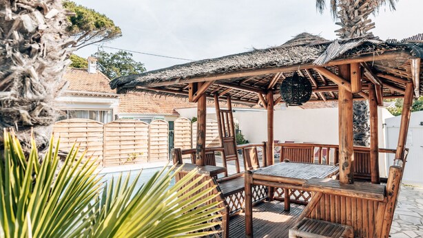 Un área de estar exterior con mesas y sillas de madera, una estructura de techo de paja y una piscina al fondo.