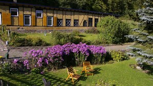 Un bâtiment à la façade en pierre et aux volets jaunes, entouré d’un jardin bien entretenu où poussent des fleurs violettes et où se trouve une chaise en bois.