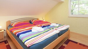Una cama individual con un edredón y almohadas a rayas de colores, una mesita de noche con una lámpara, y una ventana con vistas a árboles.