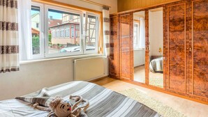 A bedroom with a bed, a window with curtains, a wooden wardrobe, and a view of a house outside.