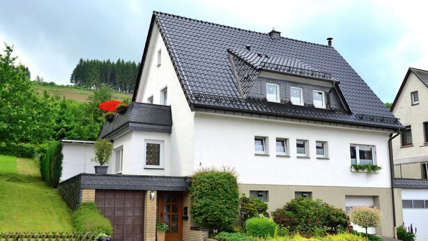 A two-story house with a dark tiled roof, a brick entrance, and a small front garden with various plants and flowers. - Graceful Apartment with Garden in Velmede near City Center (Bestwig)