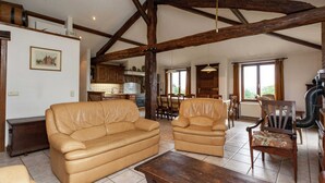 A spacious living room with a high ceiling and exposed wooden beams, featuring a leather sofa, a wooden coffee table, and a dining area with wooden chairs and a table. - Cozy Cottage, Basse-bodeux (Trois-Ponts)