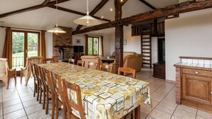 A spacious dining area with a long table, wooden chairs, and a patterned tablecloth. There are wooden beams on the ceiling, a brick fireplace, and a view of greenery outside. - Cozy Cottage, Basse-bodeux (Trois-Ponts)