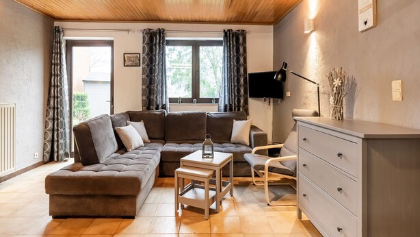 A cozy living room with a sectional sofa, a small table, a chair, and a dresser. There is a TV mounted on the wall and a window with curtains. - Cottage With Garden, Lessive (Rochefort)