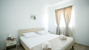 Uma cama bem arrumada com lençóis brancos e toalhas dobradas, uma mesa de cabeceira com abajur e livros, e uma janela com cortinas.